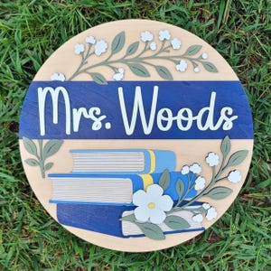 May include: A circular wooden sign featuring the text "Mrs. Woods" in white script on a dark blue horizontal band. The design includes light wood grain sections decorated with layered green leaf sprigs, white flowers, and a stack of three books.