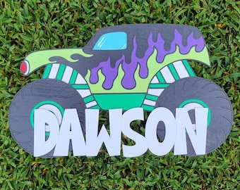 May include: A wooden monster truck cutout featuring a lime green body with purple flame graphics, a light blue window, and large black tires. The name DAWSON is displayed in large, white, block-style capital letters across the front of the truck.