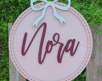 May include: A circular wooden wall sign featuring a light pink background with a dark red script name Nora in the center. The sign is topped with a light gray decorative bow and includes a stitched border detail along the edge of the circle.