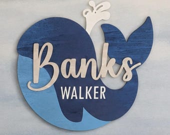 May include: A wall-mounted wooden sign shaped like a whale in shades of dark and light blue. The sign features a white water spout and the name Banks Walker in white script and block lettering layered over the whale body for a dimensional effect.