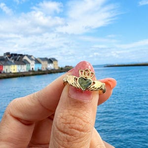 May include: A gold Claddagh ring featuring a heart-shaped green stone at the center, topped with a crown and flanked by two hands. The ring is held up against a backdrop of a blue body of water and a row of colorful buildings under a cloudy sky.