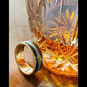 May include: A gold ring with a black and turquoise inlay. The ring is sitting on a wooden surface next to a glass of amber liquid.