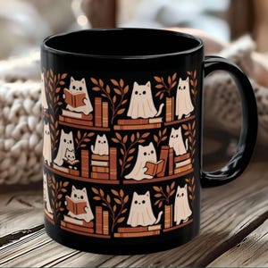 May include: A black ceramic mug featuring a repeating pattern of white ghost cats reading books and sitting on stacks of books, interspersed with orange leafy branches. The mug has a curved handle on the right side and a smooth, glossy finish.