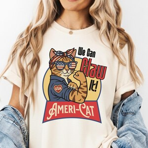 Puede incluir: Camiseta color crema con un gato de dibujos animados con gafas de sol y pañuelo, al estilo de Rosie la Remachadora. El gato flexiona su brazo, con el texto "We Can Claw It!" y "Ameri-Cat".