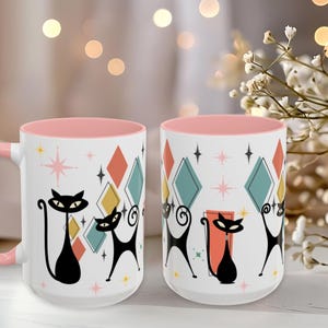 May include: Two white ceramic mugs with a pink interior and handle. The exterior features a repeating pattern of stylised black cats, colourful diamonds in teal, orange, and yellow, and small pink and yellow starburst shapes against a white background.