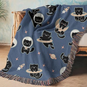 May include: A blue woven throw blanket featuring a repeating pattern of black cats in space suits, floating in space, and on planets. The blanket has a fringed edge with a mix of colors.