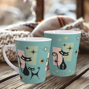 May include: Two ceramic mugs with a retro cat design. The mugs have a teal background with black cat silhouettes, starbursts, and geometric shapes in pink and green. One mug has a handle, the other is a tumbler.