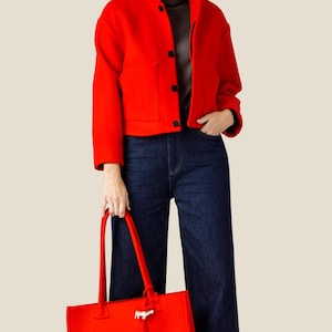 Peut inclure: Un sac fourre-tout en feutre rouge vif avec deux longues anses et une petite breloque blanche en forme d'animal attachée sur le devant. Le sac présente une structure rectangulaire et un design épuré, offrant une touche de couleur vive.
