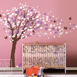 May include: A wall decal featuring a brown tree with white and purple blossoms and brown butterflies. The decal is applied to a pink wall above a crib. The text "styley walls" is in the lower left corner.