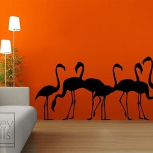 May include: Black silhouette wall decal of a flock of flamingos against an orange wall. The decal is in a living room setting with a couch and floor lamp.