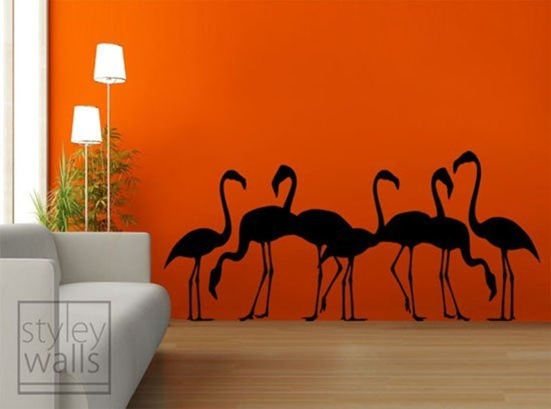May include: Black silhouette wall decal of a flock of flamingos against an orange wall. The decal is in a living room setting with a couch and floor lamp.