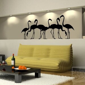 May include: Black silhouette wall decal of a flock of flamingos with their heads down, as if they are drinking water. The decal is on a wall in a living room with a yellow couch and a coffee table.