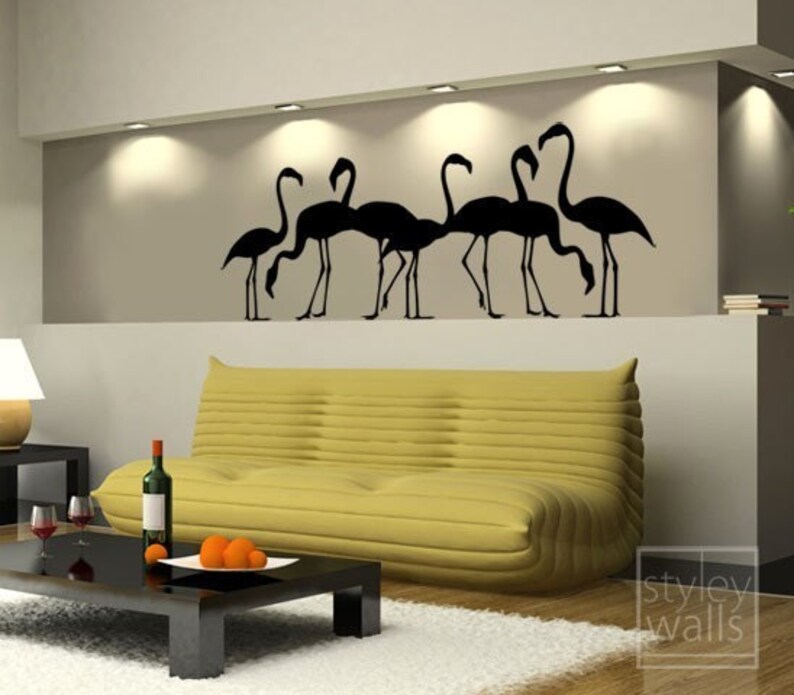 May include: Black silhouette wall decal of a flock of flamingos with their heads down, as if they are drinking water. The decal is on a wall in a living room with a yellow couch and a coffee table.