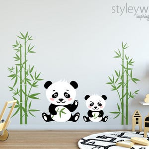 May include: A set of two panda bear wall decals with bamboo stalks. The larger panda is holding a bamboo leaf. The smaller panda is holding a bamboo stalk. The decals are white and black with pink accents. The background is a light grey wall.