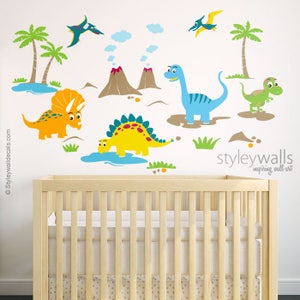May include: A set of colorful dinosaur wall decals featuring a blue dinosaur, a yellow dinosaur, an orange dinosaur, a green dinosaur, a flying dinosaur, palm trees, and volcanoes. The decals are on a white wall with a wooden crib in the foreground. The text "styleywalls inspiring wall art" is in the bottom right corner of the image.