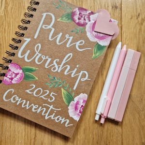 May include: A brown notebook with a white painted floral design and the words "Pure Worship 2025 Convention". A pink heart-shaped paper clip is attached to the notebook. Three pink and white pens are lying on a wooden surface.