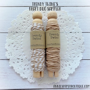 May include: Two spools of twine on wooden dowels. The twine on the left is white and brown, and the twine on the right is brown. Both spools have labels that say "Trendy Twine" and "AnniesPaperBoutique.com".