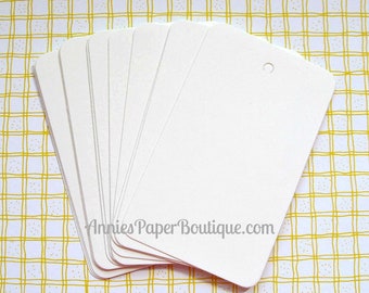 May include: A stack of ten blank white gift tags with a hole punched in the top corner. The tags are arranged in a fan shape and have a website address printed on the bottom: AnniesPaperBoutique.com