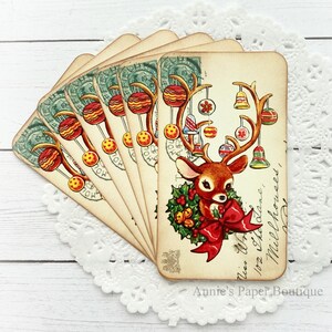 May include: A stack of vintage-style Christmas cards featuring a reindeer with festive ornaments on its antlers and a red bow. The cards have a distressed, aged appearance with a green postage stamp design and handwritten text.