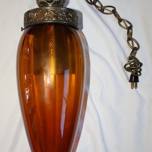 May include: An amber glass pendant light with a decorative brass top and chain. The light fixture has a teardrop shape and a brass chain for hanging. The plug is black. This vintage-style light fixture is suitable for home decor.