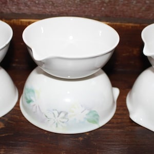 May include: Three white ceramic bowls with a pouring spout, stacked on top of matching bases. One base has a floral design in pastel colors. The bowls are on a dark wooden surface.