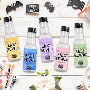 Puede incluir: Cinco botellas de vidrio con tapas negras y etiquetas coloridas que dicen "Baby Brewing Potion" con un gráfico de caldero. Decoraciones temáticas de Halloween, incluyendo murciélagos, una calabaza y caramelos, rodean las botellas.