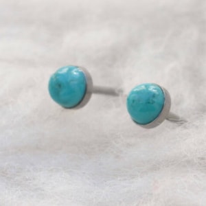May include: A pair of turquoise stud earrings. Each earring features a round, polished turquoise stone set in a silver-colored bezel. The earrings are displayed on a white, textured surface.