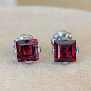 May include: A pair of square-cut, deep red gemstone stud earrings. The gemstones are set in a silver-colored metal setting. The earrings are displayed on a neutral-toned surface.