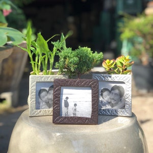May include: Three silver picture frames with different photos inside. The frames are decorated with a leaf pattern. The frames are sitting on a light gray surface.