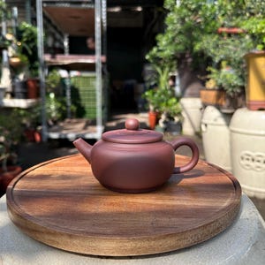 May include: A small, dark red clay teapot with a lid and a handle. The teapot is sitting on a wooden circular tray.