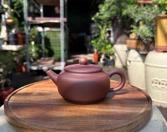 May include: A small, dark red clay teapot with a lid and a handle. The teapot is sitting on a wooden circular tray.
