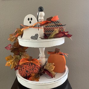 May include: A white two-tiered tray with a white ghost, orange and black pumpkins, and autumn leaves. The tray is on a dark brown table.