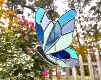 May include: A stained glass butterfly garden stake with a copper stem. The butterfly features a mosaic design in shades of blue, teal, and gray. The wings are outlined in black. The garden stake is displayed outdoors near a fence and flowering plants.