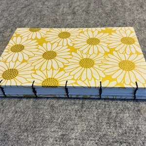 May include: A handmade journal with a yellow floral daisy pattern cover. The journal is bound with black thread and has a rectangular shape. The pages are visible along the spine.