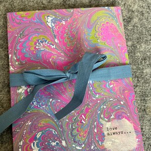 May include: A notebook with a colorful marbled design in pink, blue, green, and white, tied with a blue ribbon. The notebook has the words "love always..." printed on it.