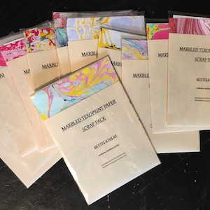 May include: A set of marbled paper scraps in various colors, including pink, yellow, blue, and white. The paper is packaged in clear plastic sleeves and labeled "Marbled Texoprint Paper Scrap Pack" with the artist's name and website.