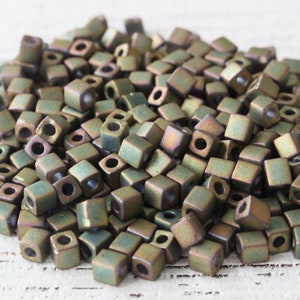 May include: A pile of small, square, metallic beads in a muted green colour with a slight iridescent sheen.