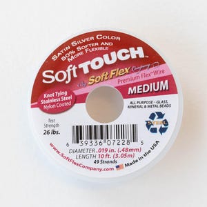 Op de afbeelding: Een spoel zilverkleurige Soft Touch Soft Flex Premium Flex draad voor kralen. Het is middelgroot, 3,05 meter lang en heeft een diameter van 0,48 mm. De spoel is gelabeld "All Purpose - Glas, Mineralen en Metalen kralen".
