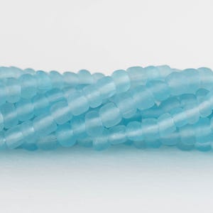 Puede incluir: Primer plano de un hilo de cuentas cilíndricas de color azul claro. Las cuentas tienen un aspecto ligeramente esmerilado y están ensartadas, creando una superficie texturizada. Las cuentas son de un color azul pastel suave.