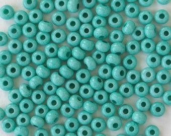 Può includere: Una collezione di perline di legno color turchese. Ogni perlina è rotonda con un foro al centro. Le perline sono sparse su una superficie chiara, creando un motivo strutturato.