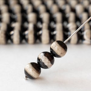 May include: Close-up of three round beads with a black and white striped pattern, strung on a silver needle. The background features a blurred array of similar beads, suggesting a jewelry-making theme.