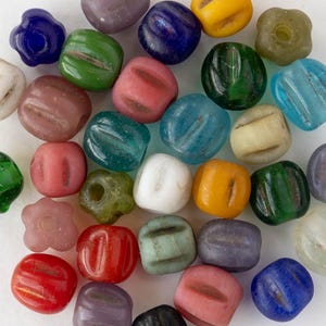 May include: A collection of colorful glass beads in various shapes and sizes. The beads are in shades of blue, green, pink, yellow, and white. Some beads have a ribbed design, while others are flower-shaped or round. The beads are likely for jewelry making.