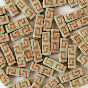 May include: A collection of light green glass beads with a copper-colored Greek key design. The beads are rectangular in shape and have a distressed, aged look.