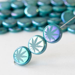 May include: Iridescent teal glass beads with a cannabis leaf design. The beads are round and have a flat back. The beads are strung on a silver wire. The background is a blurred collection of similar beads.