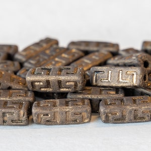 May include: A pile of dark brown ceramic beads with a gold Greek key pattern. The beads are rectangular in shape and have a slightly rough texture.