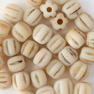 May include: An overhead shot of a collection of off-white beads. The beads are primarily barrel-shaped with three dark brown stripes. Two flower-shaped beads are also present. These beads are ideal for jewelry making and crafting.
