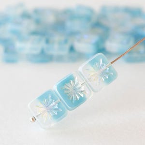 May include: A strand of square, light blue and white glass beads with a starburst design. The beads are strung on a thin wire, with a silver bead at the end. The background features more out-of-focus beads.