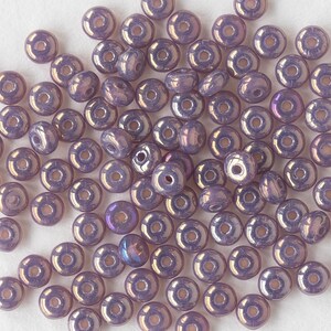 Pu&ograve; includere: Un grande gruppo di piccole perline di vetro rotonde e iridescenti di colore viola. Le perline hanno un foro al centro e sono sparse su una superficie bianca.