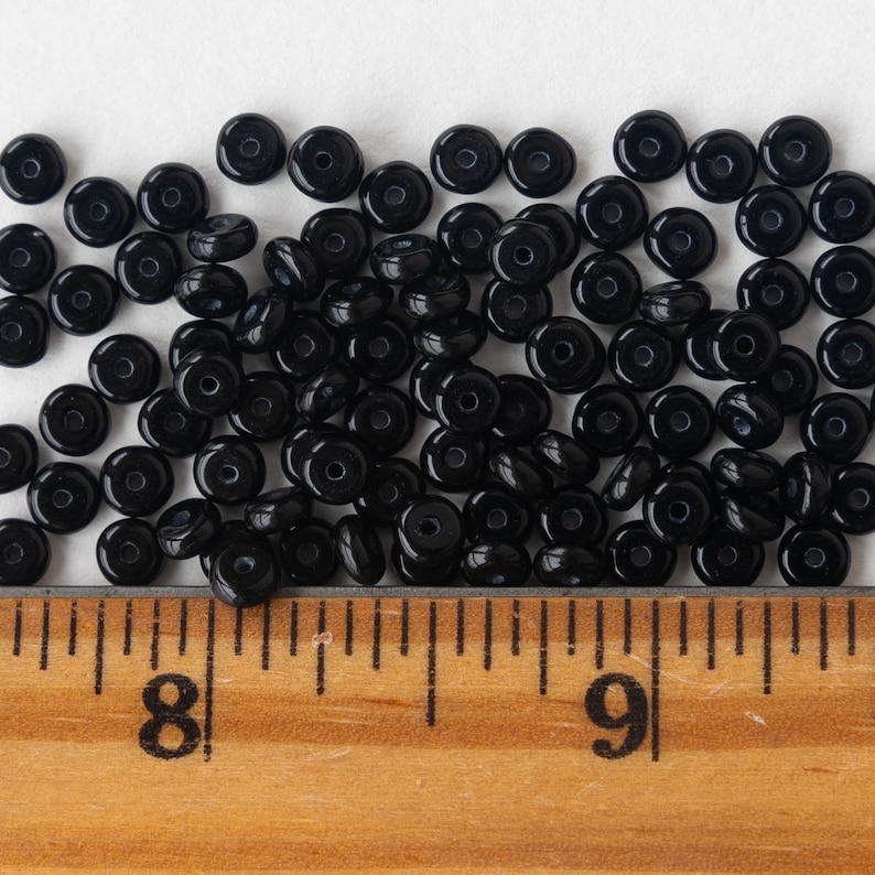 May include: A close-up of a pile of black glass beads, measured against a wooden ruler. The beads are round with a small hole in the center.
