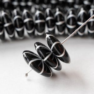 May include: Close-up of black and white oval beads strung on a silver wire. The beads have a glossy finish with a white stripe design. The background shows more beads, suggesting a craft supply or jewelry component.